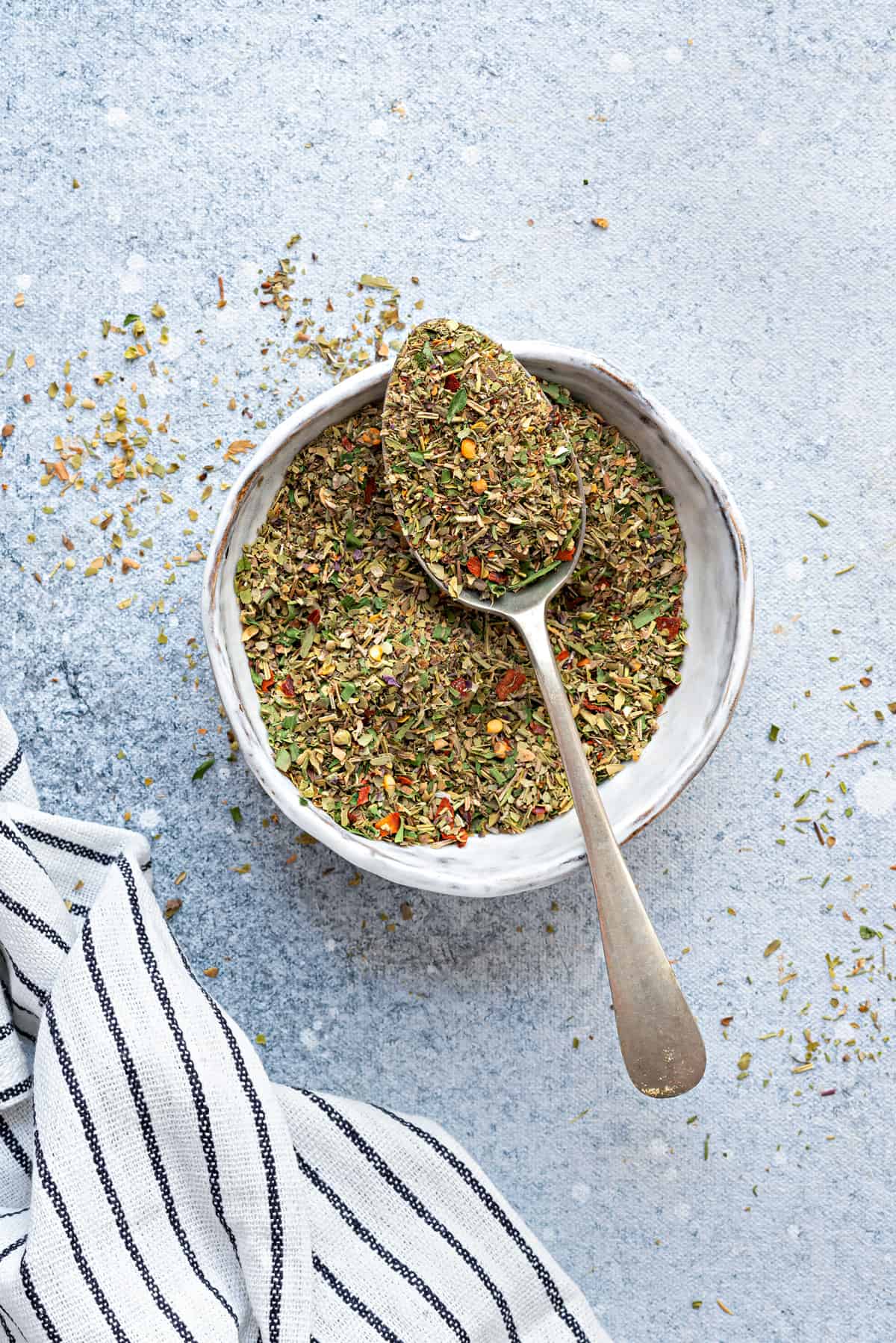 a white bowl of Italian Seasoning mix with a spoon on blue table.