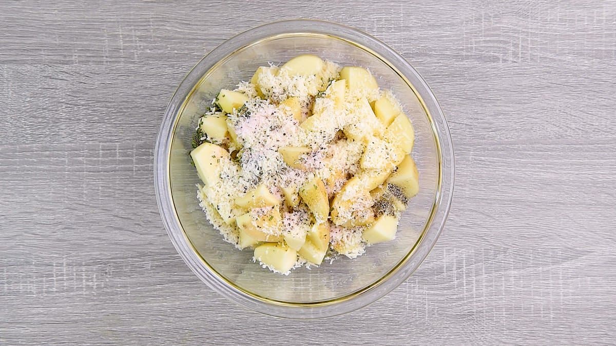 Cubed potatoes, olive oil, garlic, chopped rosemary, parmesan, salt and pepper in a round glass bowl.