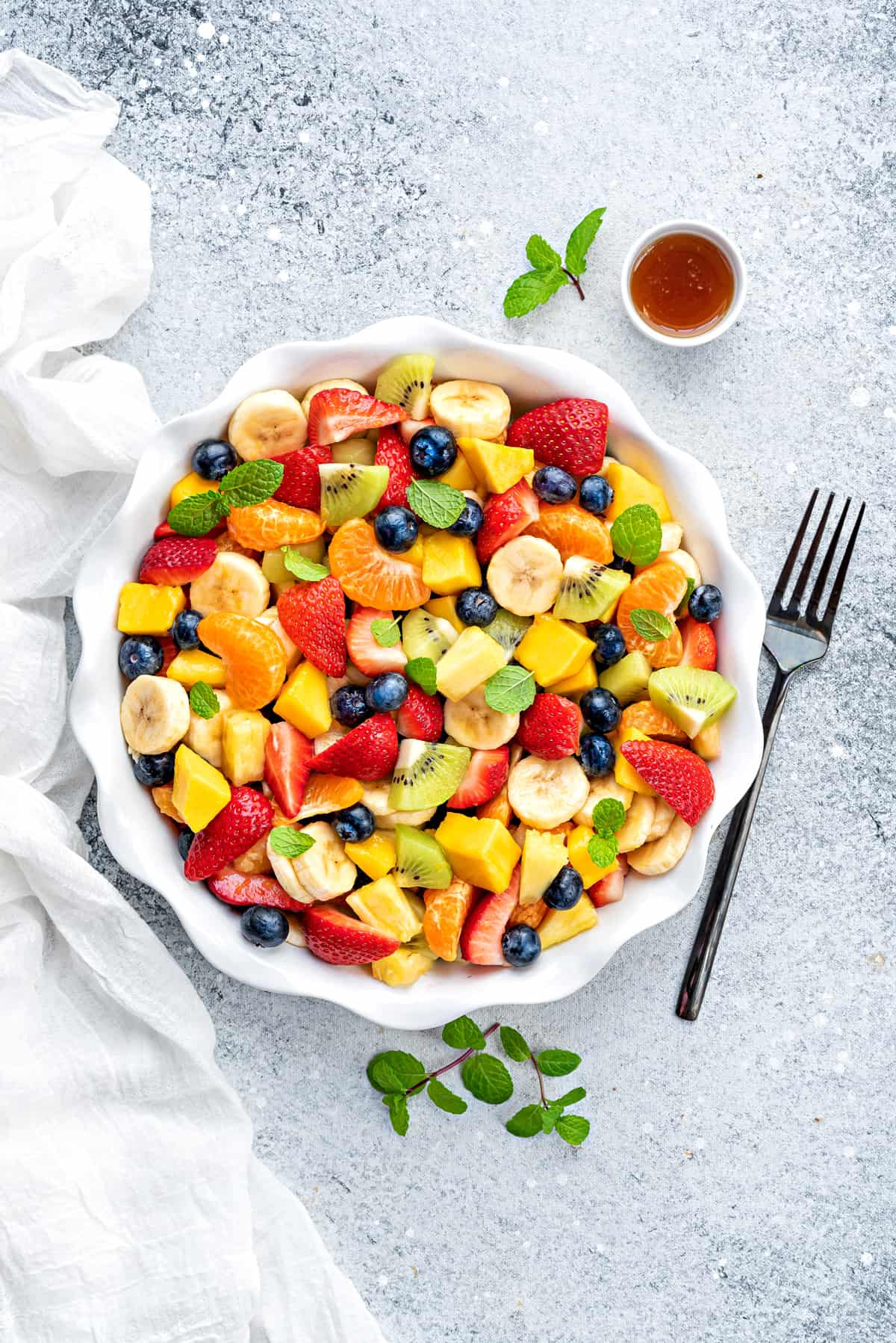 Fresh fruit salad bowl with a fork and a salad dressing bowl kept on white table.