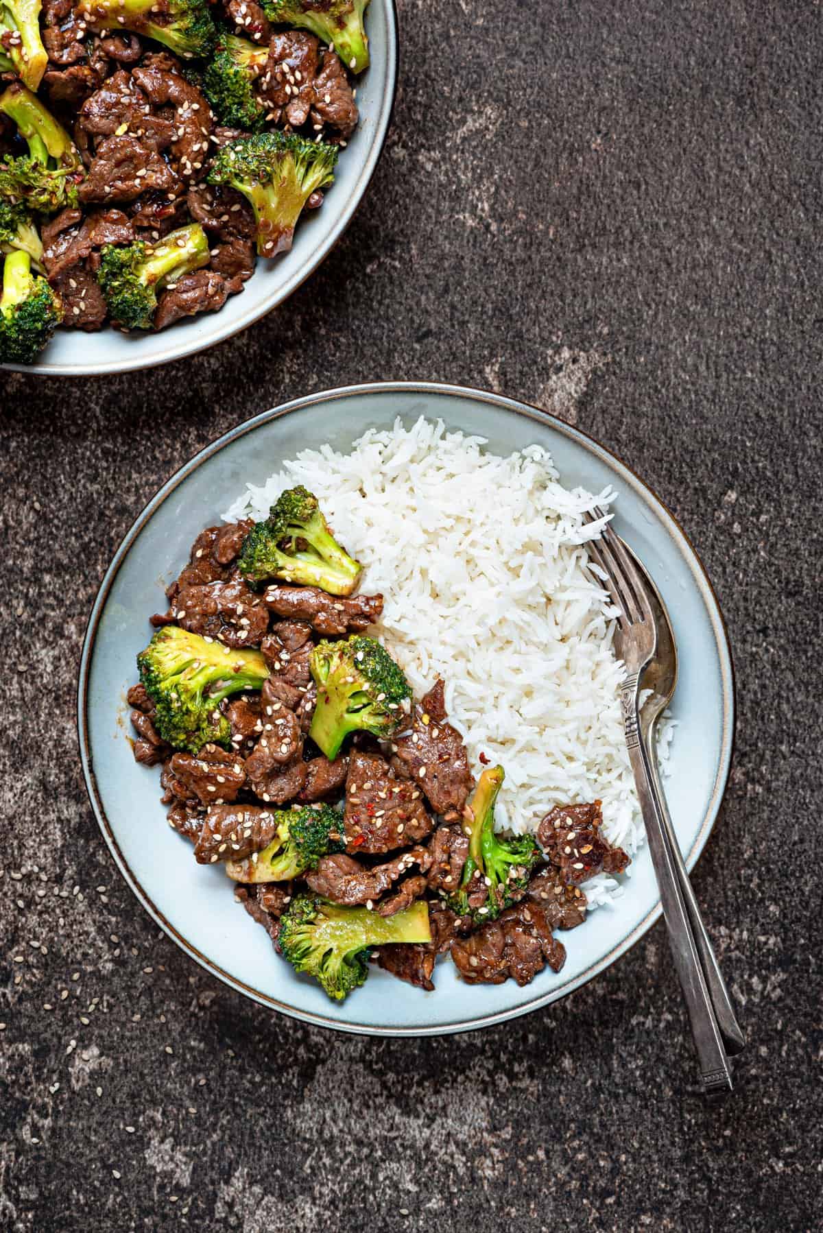 broccoli beef served in a gray bowl with a side of white rice, topped with sesame seeds and red chili flakes.