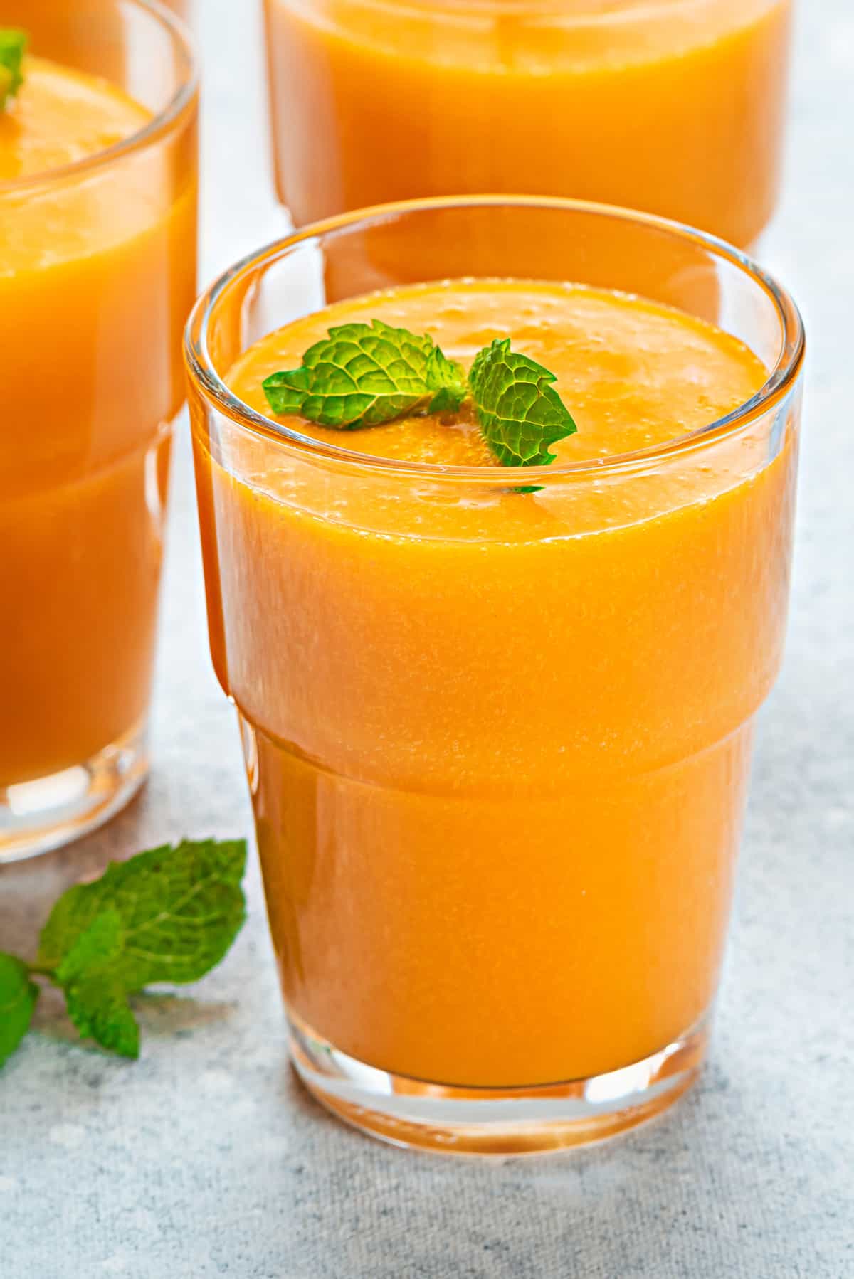 Fresh homemade mango juice in three serving glasses kept on a grey table.