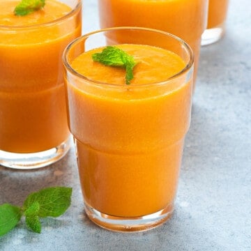 Fresh homemade mango juice in three serving glasses kept on a grey table.