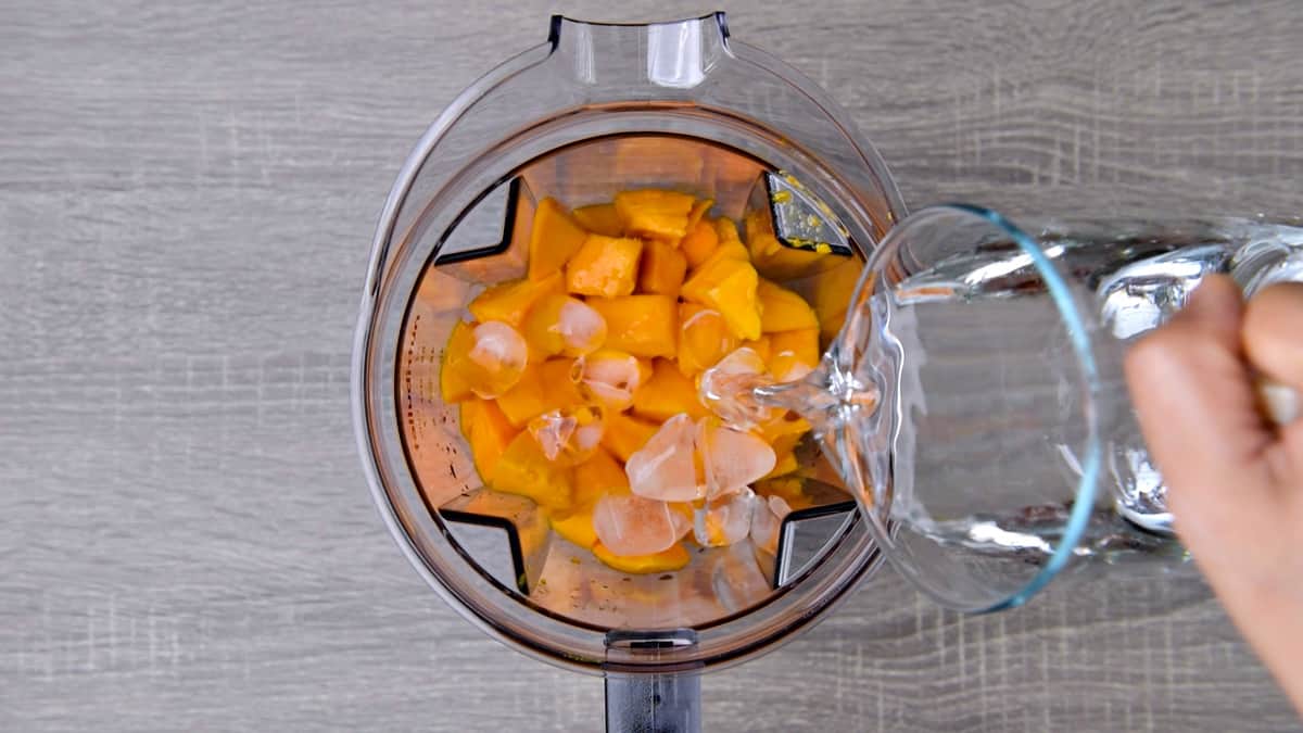Adding diced mangoes, ice cubes and water in a blender jar.