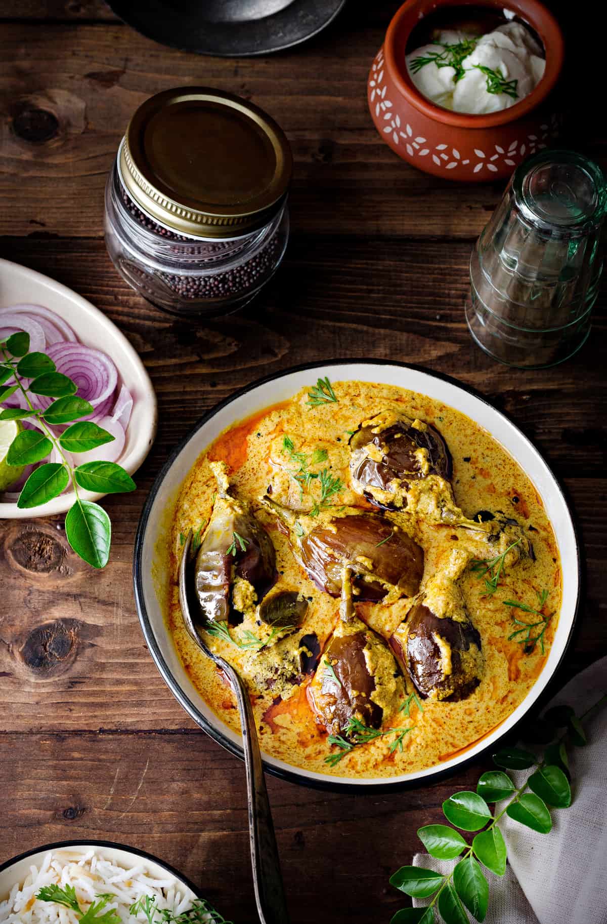 aerial shot of bagara baingan curry served in bowl with a spoon, rice and onion rings.