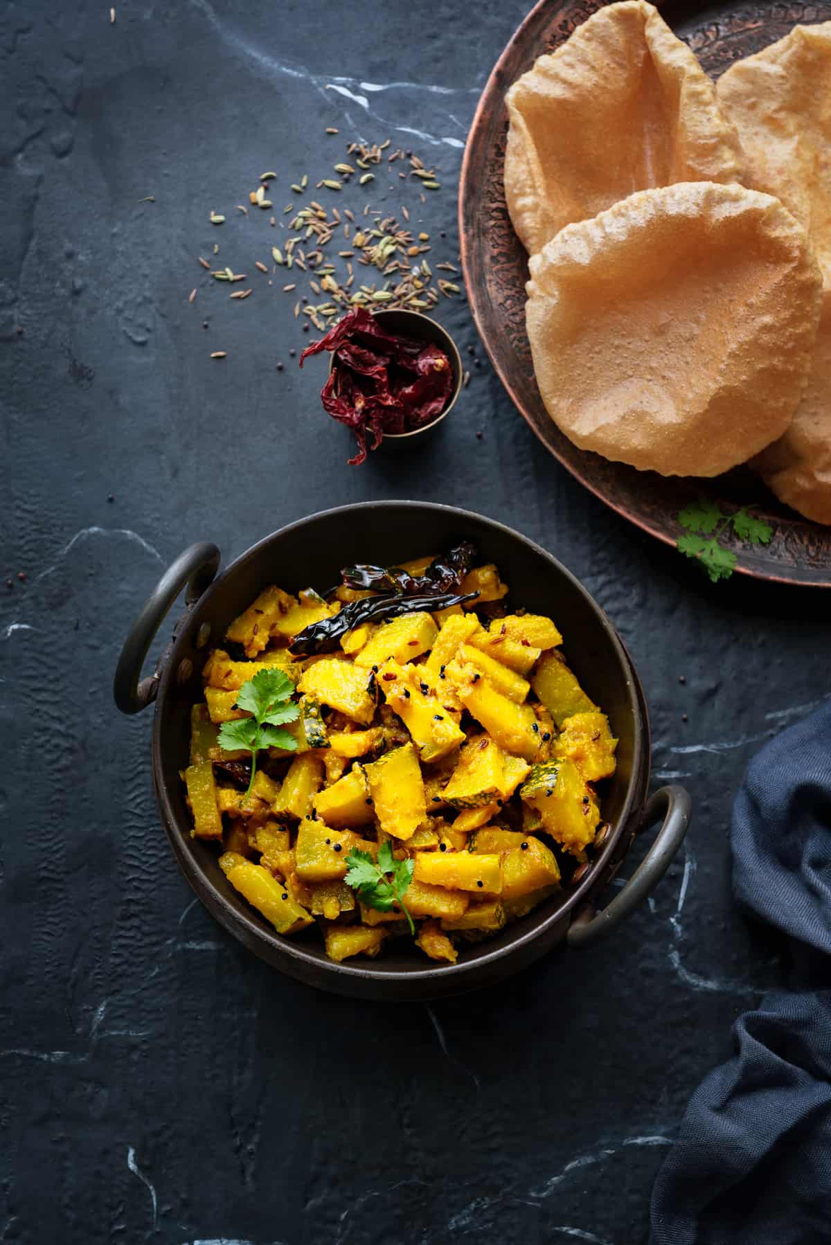 Kaddu ki Sabji served in black kadai with puris on a plate, some spices scattered around.