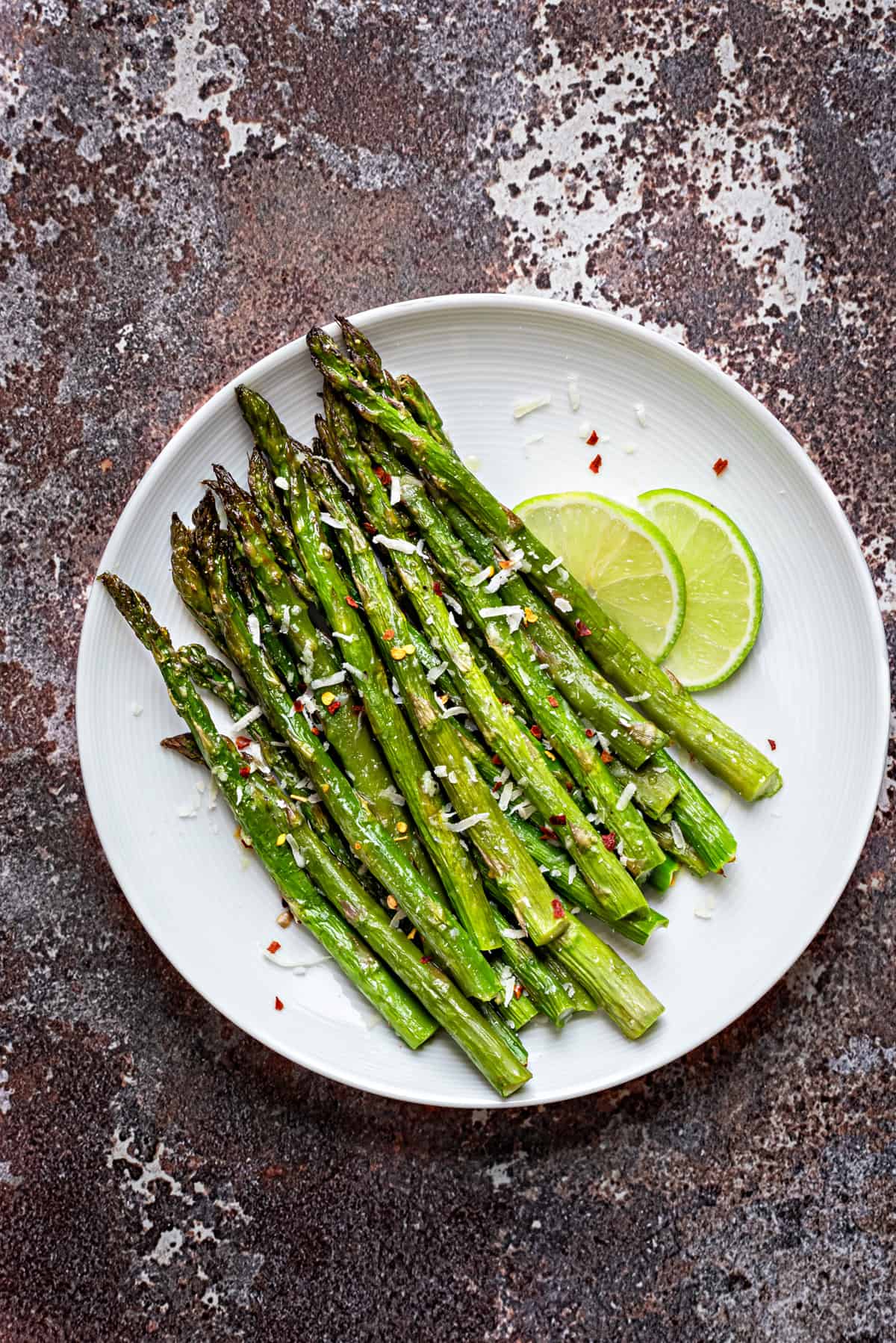 Oven roasted asparagus placed on a white plate and topped with grated parmesan, and chili flakes.