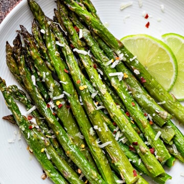 Oven roasted asparagus placed on a white plate and topped with grated parmesan, and chili flakes.