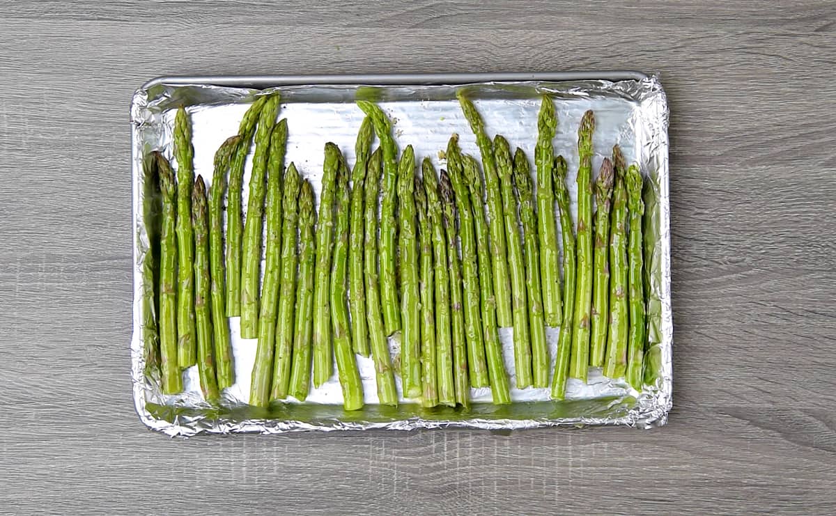 Raw asparagus lined up on a baking sheet, coated in olive oil. 