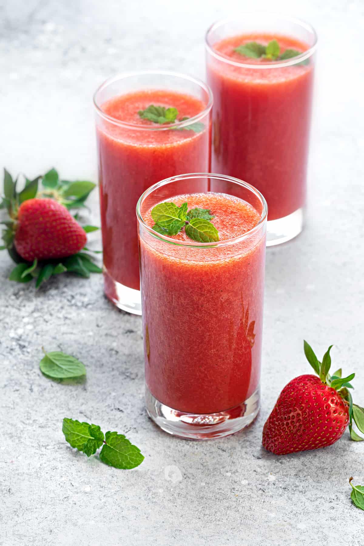 Three tall glasses with pure strawberry juice on a white table, few fresh strawberries scattered around.