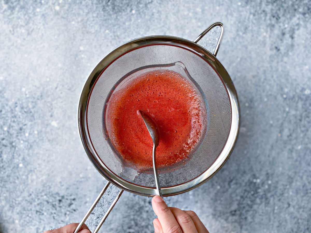 Straining the blended juice of strawberries in bowl using a fine mesh sieve and a spoon.