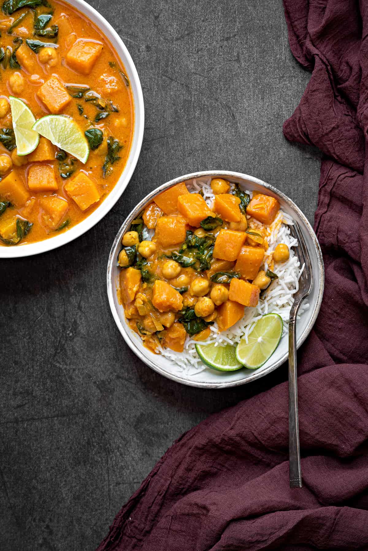 A bowl of white rice topped with butternut squash curry, lime wedges and a fork.