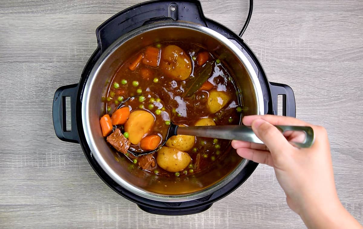 A hand holding a ladle with pressure cooked beef stew with potato and carrots over the pot.