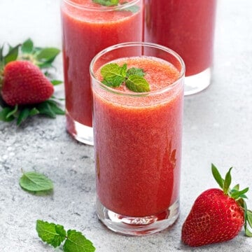 Fresh strawberry juice in a glass, two glasses with juice, mint, and strawberries in the background.