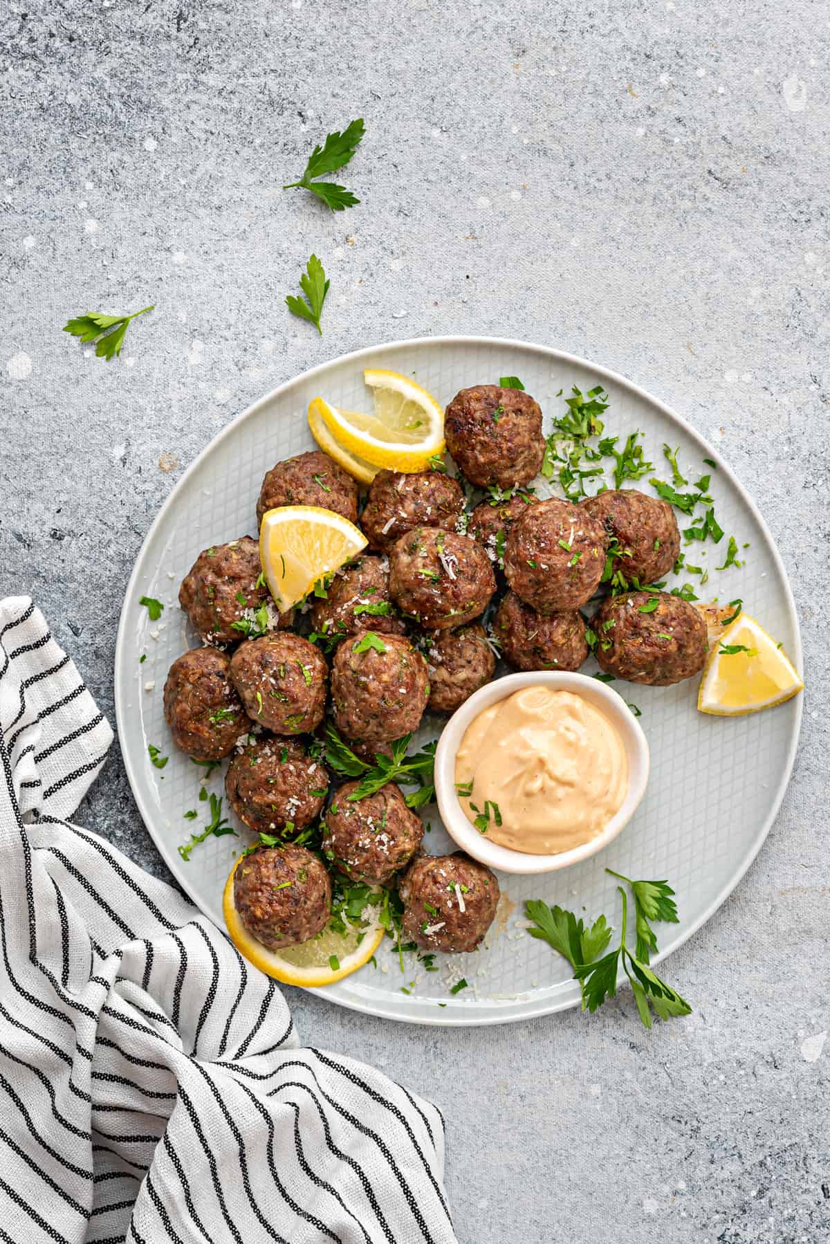 Plate with homemade baked meatballs, sprinkled with fresh parsley, lemon slices and dipping sauce in bowl.