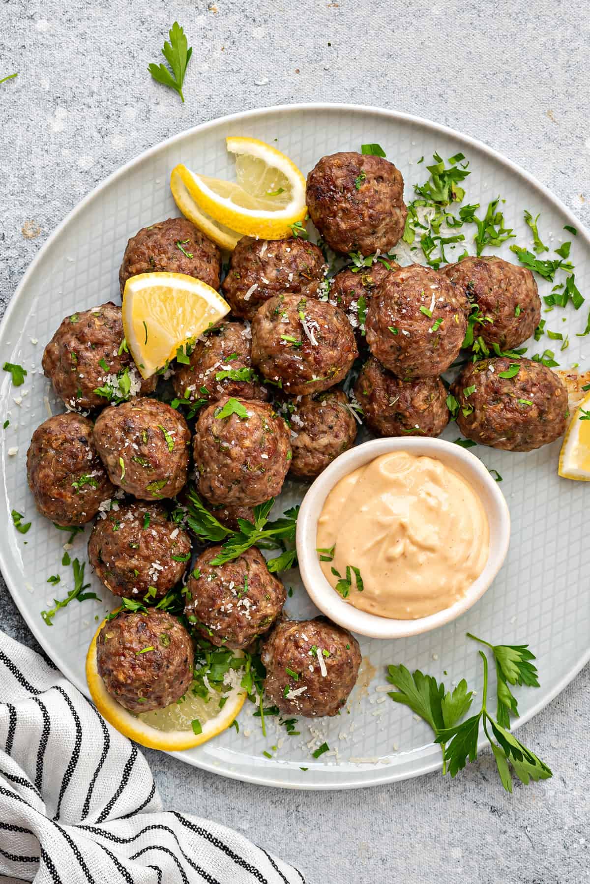 Close-up of homemade beef meatballs, sprinkled with fresh parsley, lemon slices and aioli dipping sauce in small bowl.