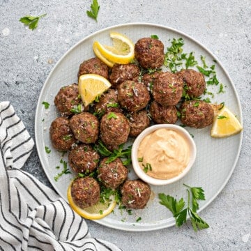 Plate with homemade beef baked meatballs, sprinkled with fresh parsley, lemon slices and dipping sauce in bowl.