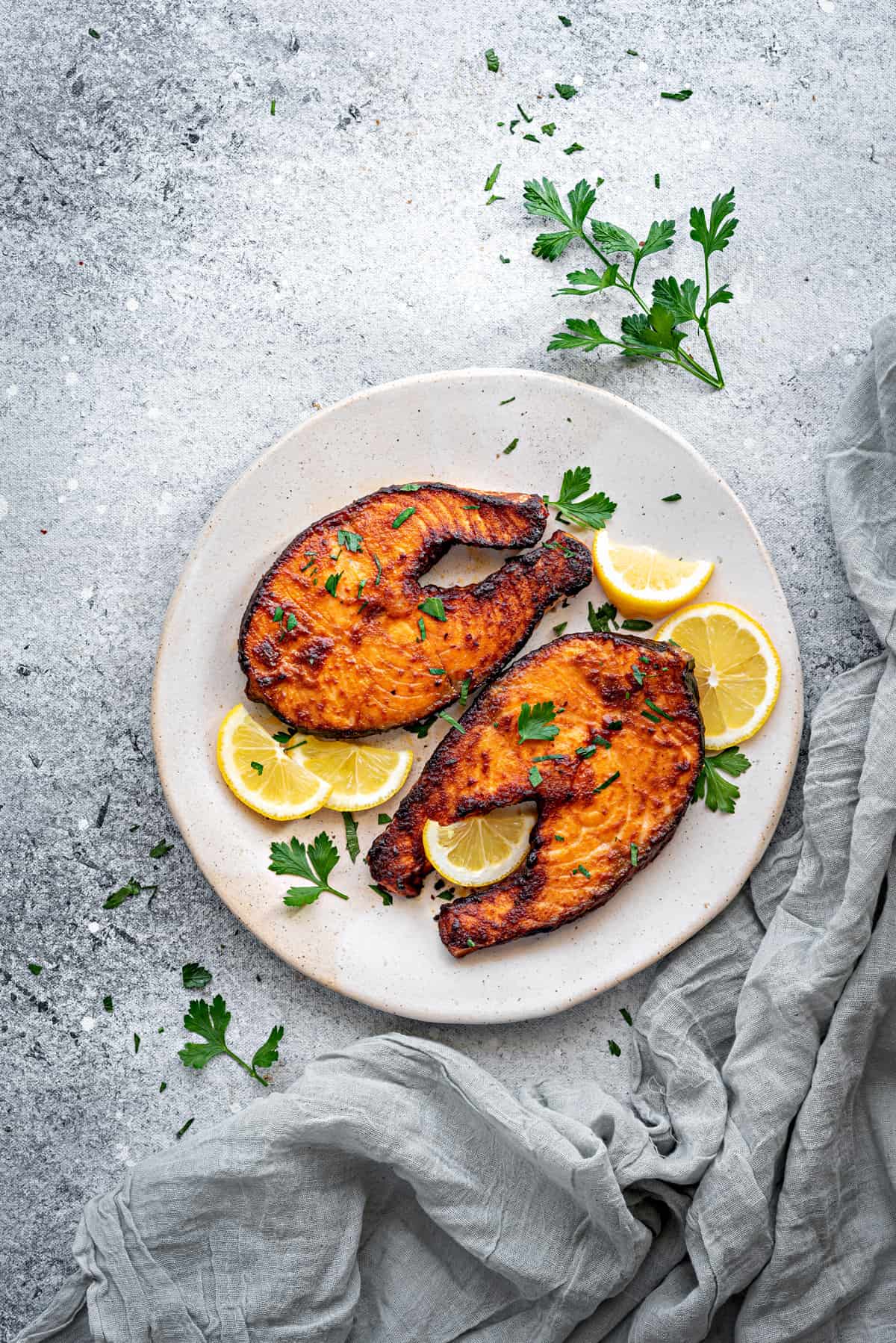 Top down view two crispy air fried salmon steaks on a white plate with slices of lemon and parsley.