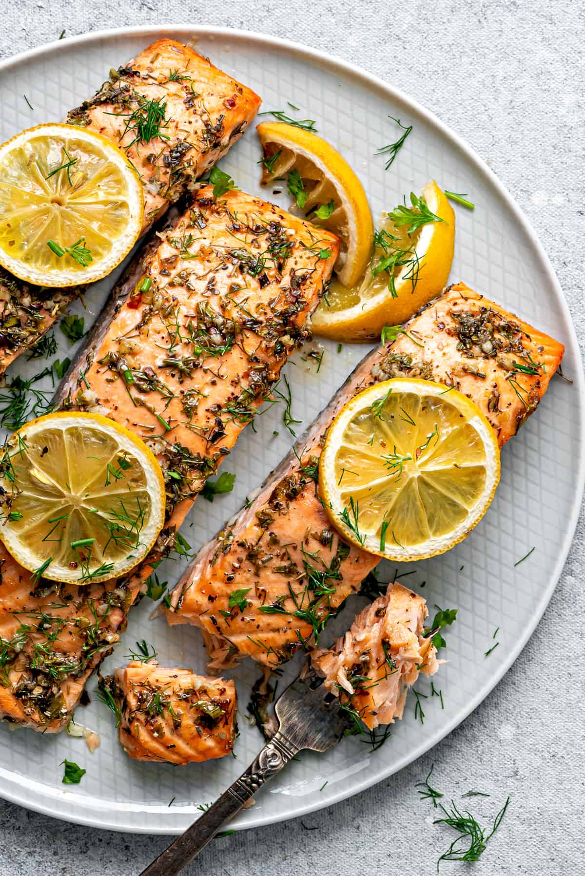 Top down three portions of oven baked salmon topped with herbs and lemon slices, on a plate.