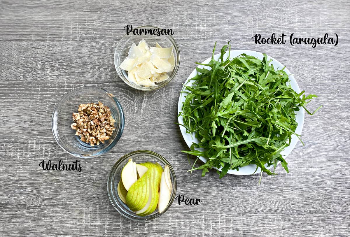 Pear salad base ingredients in individual bowls: Arugula, pear, walnuts, and shaved parmesan cheese.