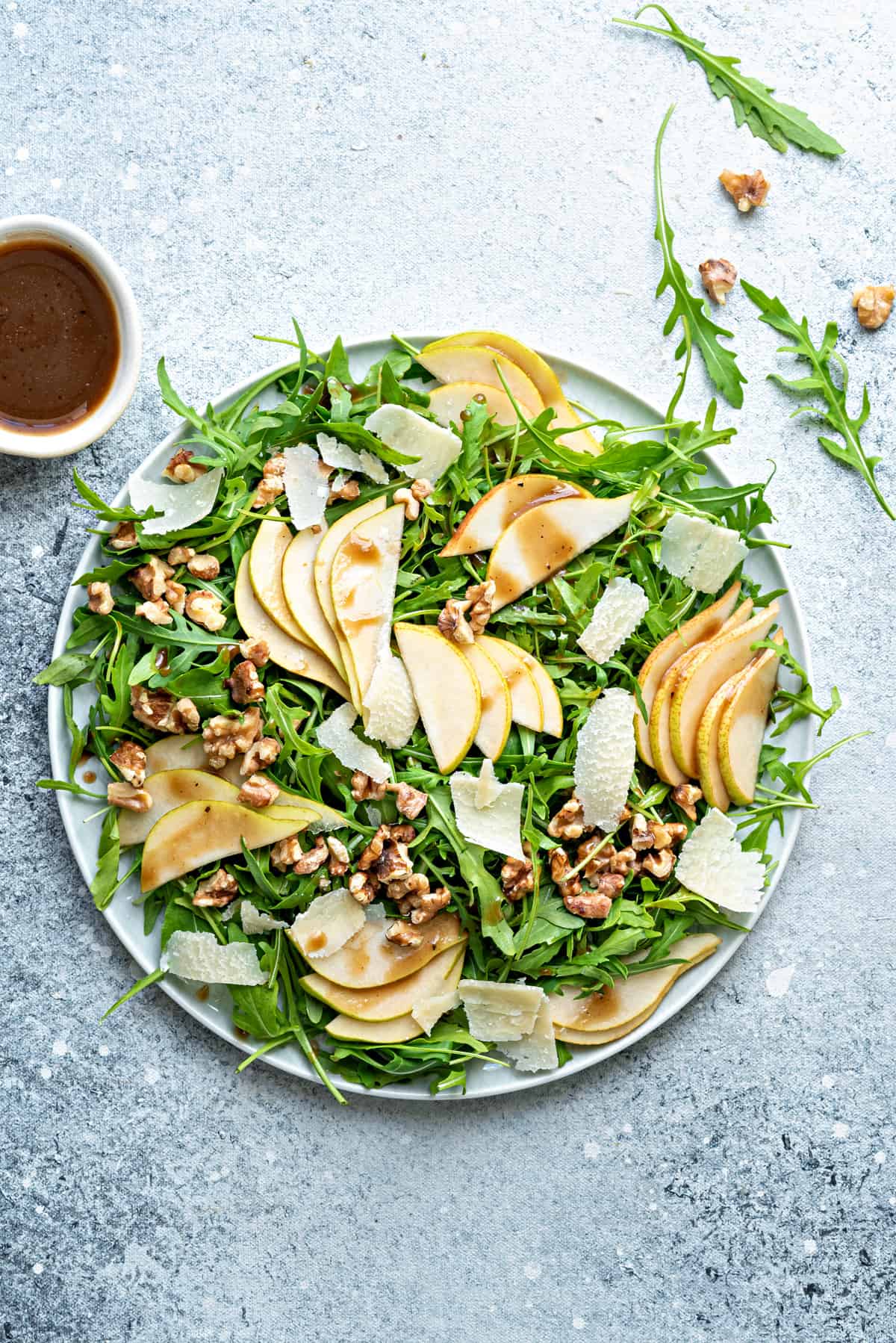 Top down large plate of pear and rocket salad with balsamic vinaigrette in bowl on side.