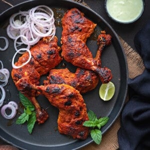 Top down close up Tandoori Chicken on balck round tray with chutney in bowl on side.