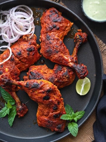 Top down close up Tandoori Chicken on balck round tray with chutney in bowl on side.