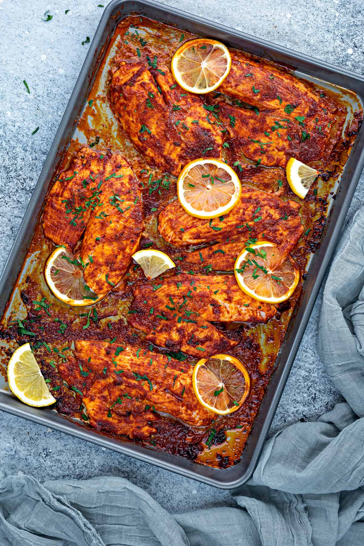 Top down view sheet pan with 6 chili lemon baked tilapia fillets with fresh lemon slices.