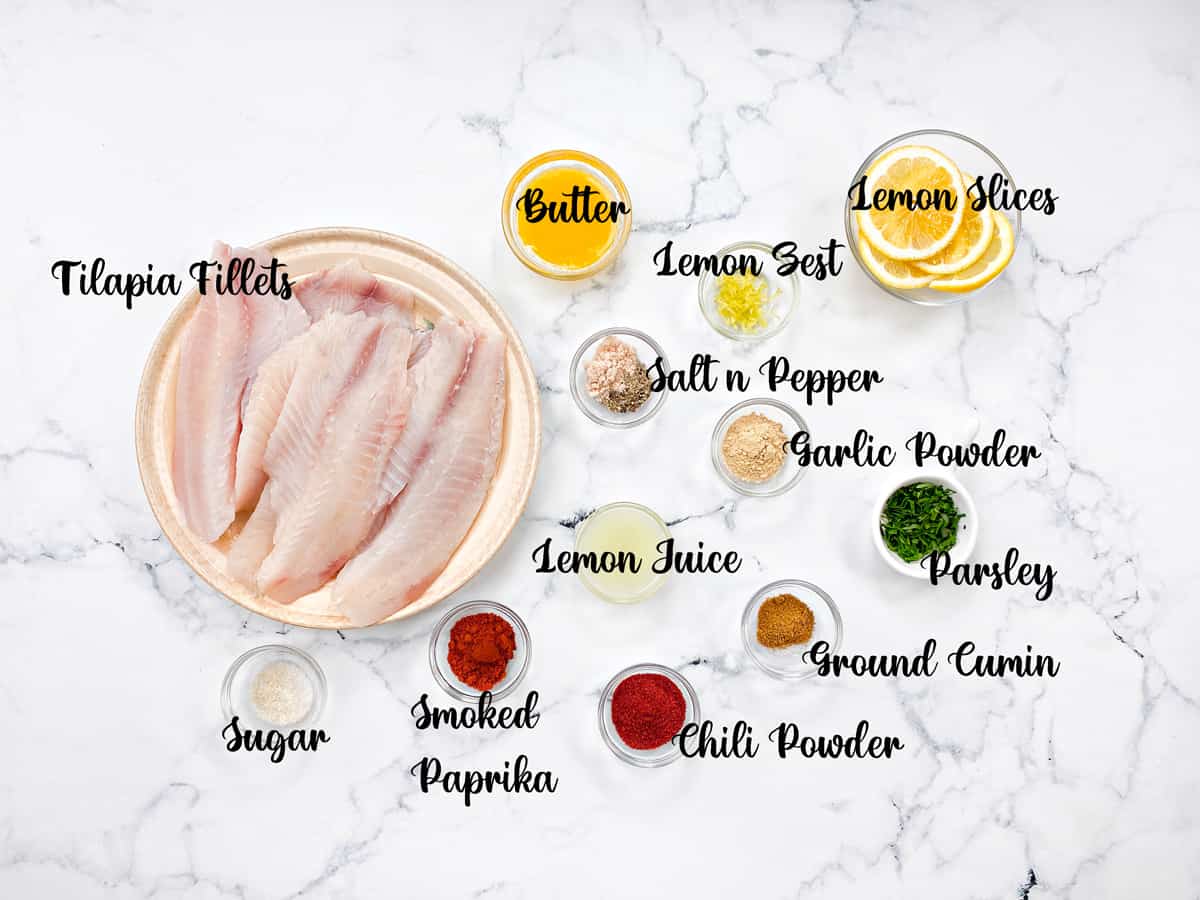 Recipe ingredients on a marble background. Each ingredient is in its own glass bowl: Tilapia fillets, melted butter, lemon slices, lemon zest, salt and pepper, garlic power, chopped parsley, lemon juice, ground cumin, chili powder, smoked paprika, and sugar.