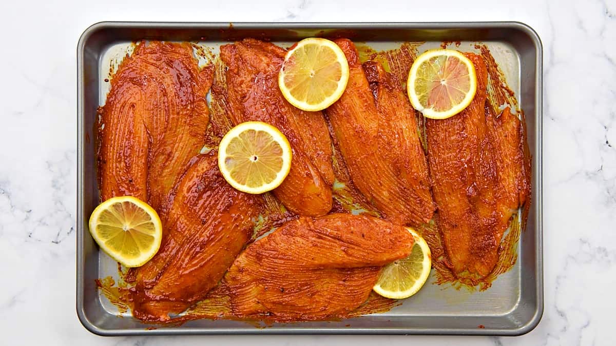 6 raw pieces of tilapia on a baking sheet with marinade and lemon slices.