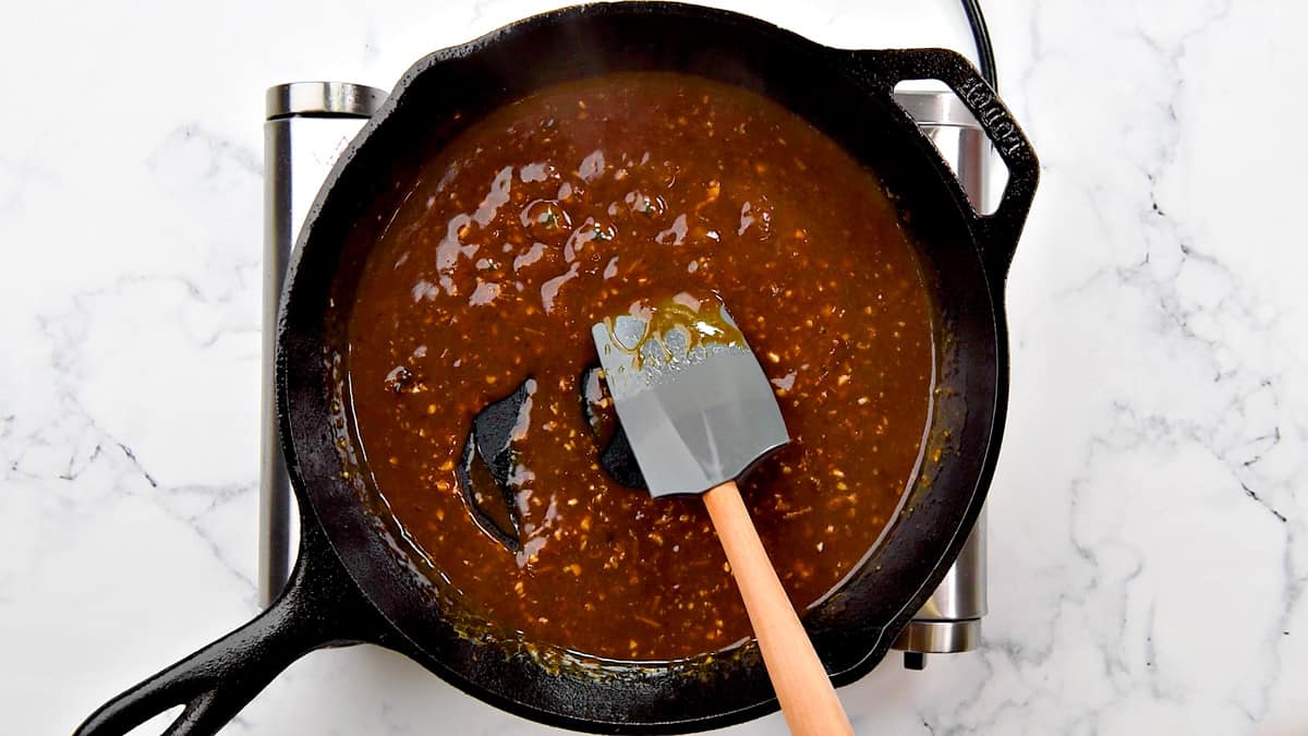 Gray spatula stirring the Asian sauce in a cast iron skillet.