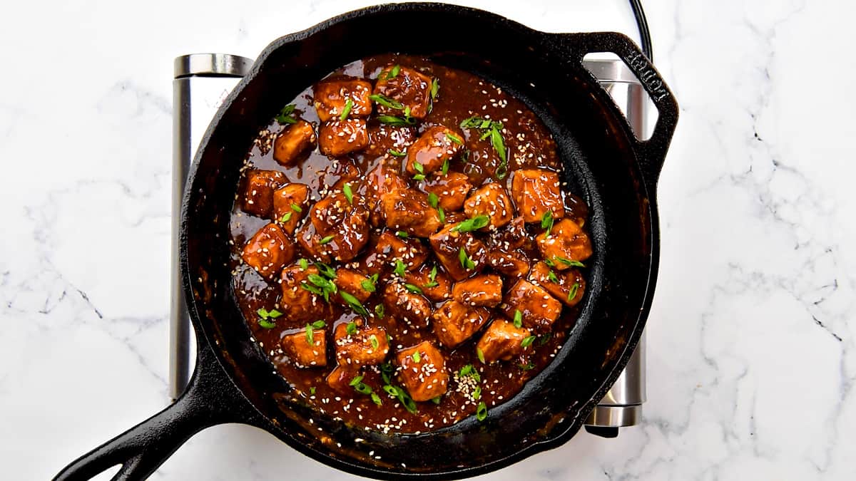 Bites of Asian salmon in a dark sauce in cast iron skillet garnished with sesame seeds and green scallions.