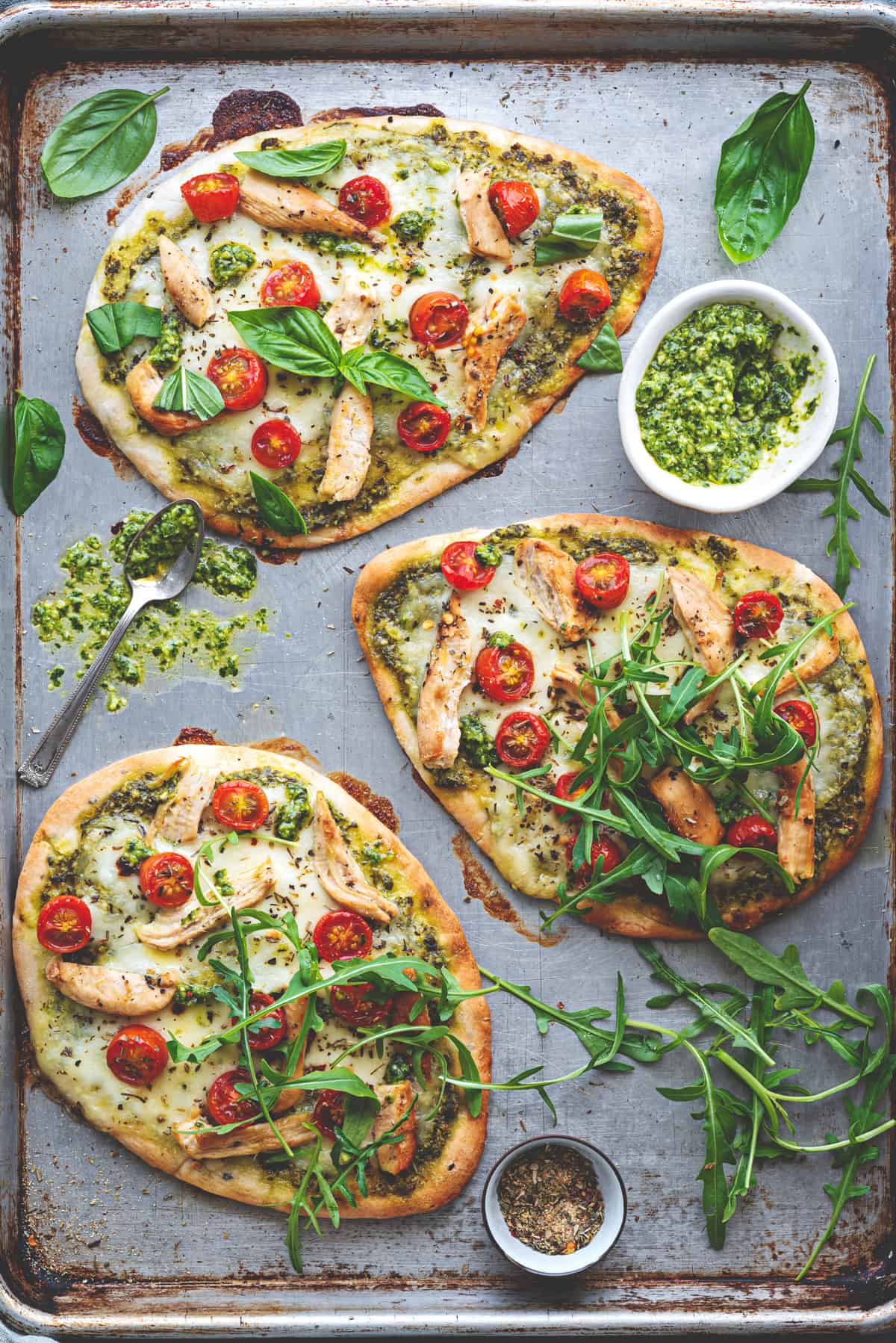 Three naan pizzas on a baking sheet, with fresh herbs and a white ramekin of pesto to the side.