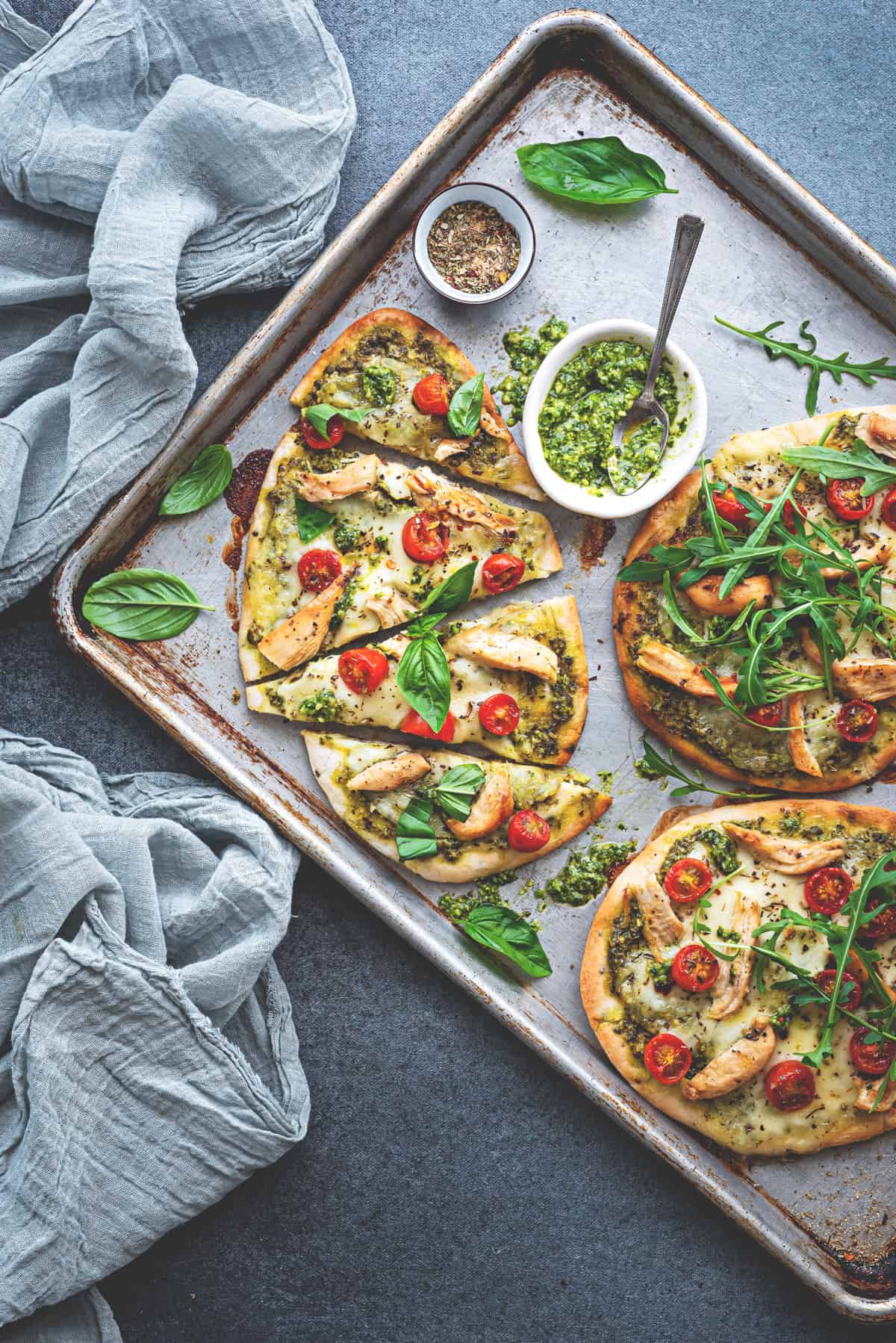 Naan pizzas cut into triangles on a baking sheet. Sprinkles of fresh basil leaves all around, and small white ramekin of pesto to the side.