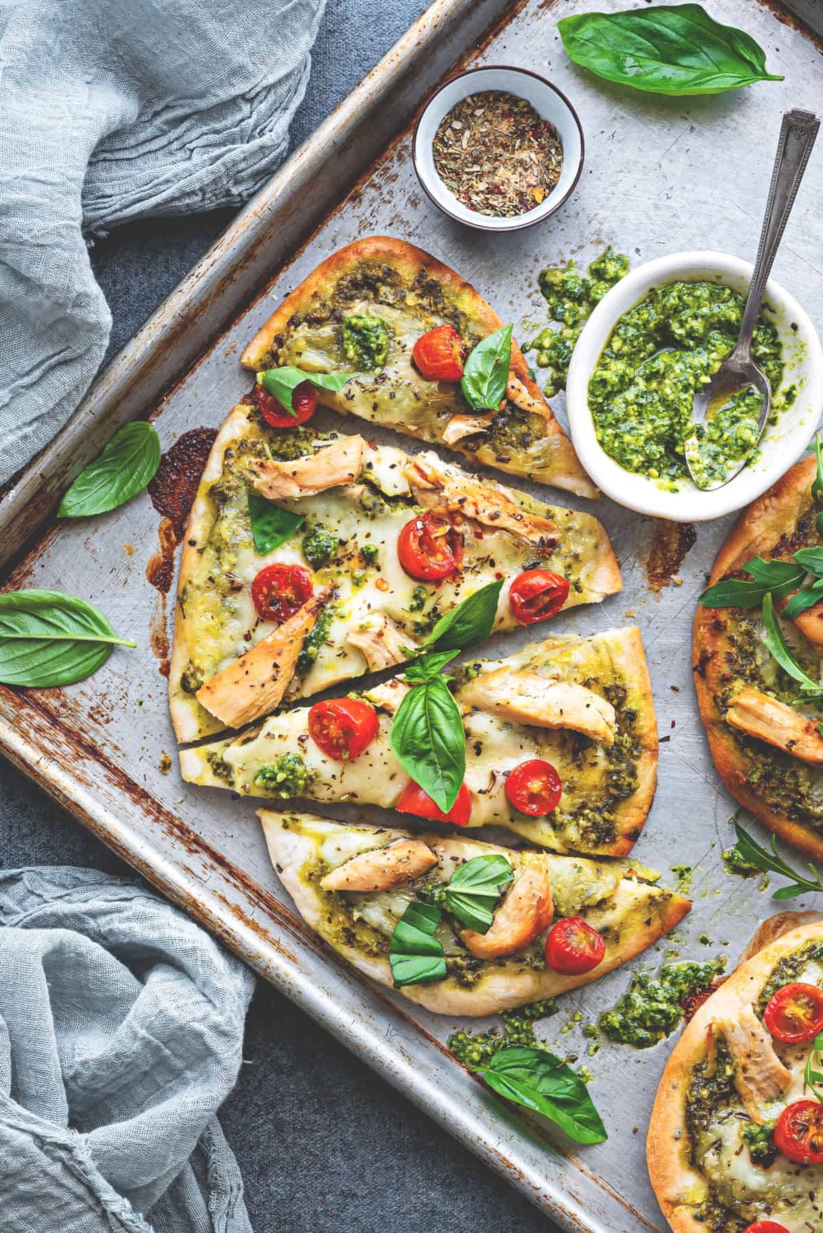 Close view naan pizzas cut into triangles on a baking sheet with fresh basil around and pesto in a small bowl.
