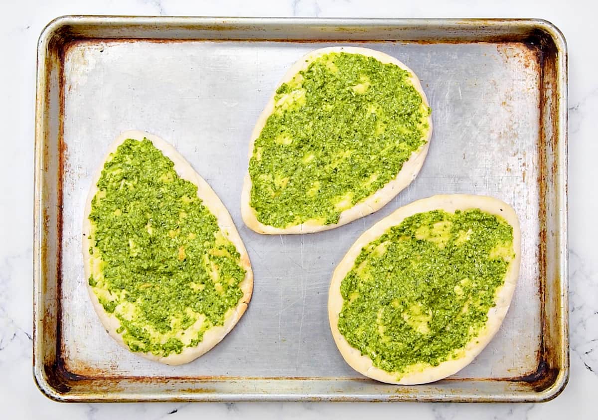 3 pieces of naan on a baking sheet. Each is covered with a layer of green basil pesto.