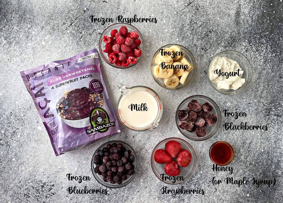 Top down view of ingredients: Package of acai puree, frozen raspberries, blueberries, and strawberries, yogurt, milk, and honey.