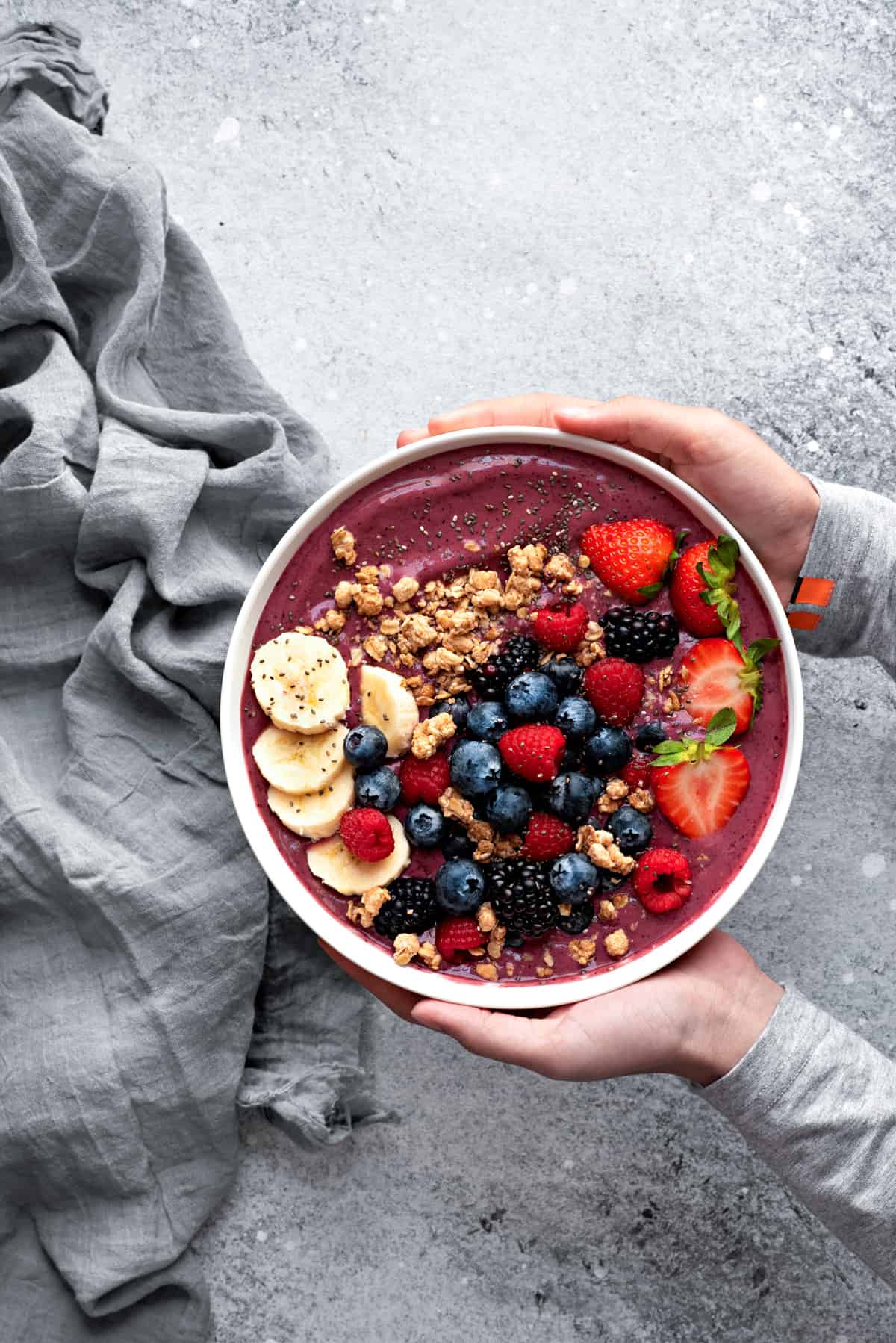 Two hands holding a large white bowl with blended acai berry smoothie, topped with slices of bananas, granola, fresh blueberries, strawberries, raspberries, and seeds.