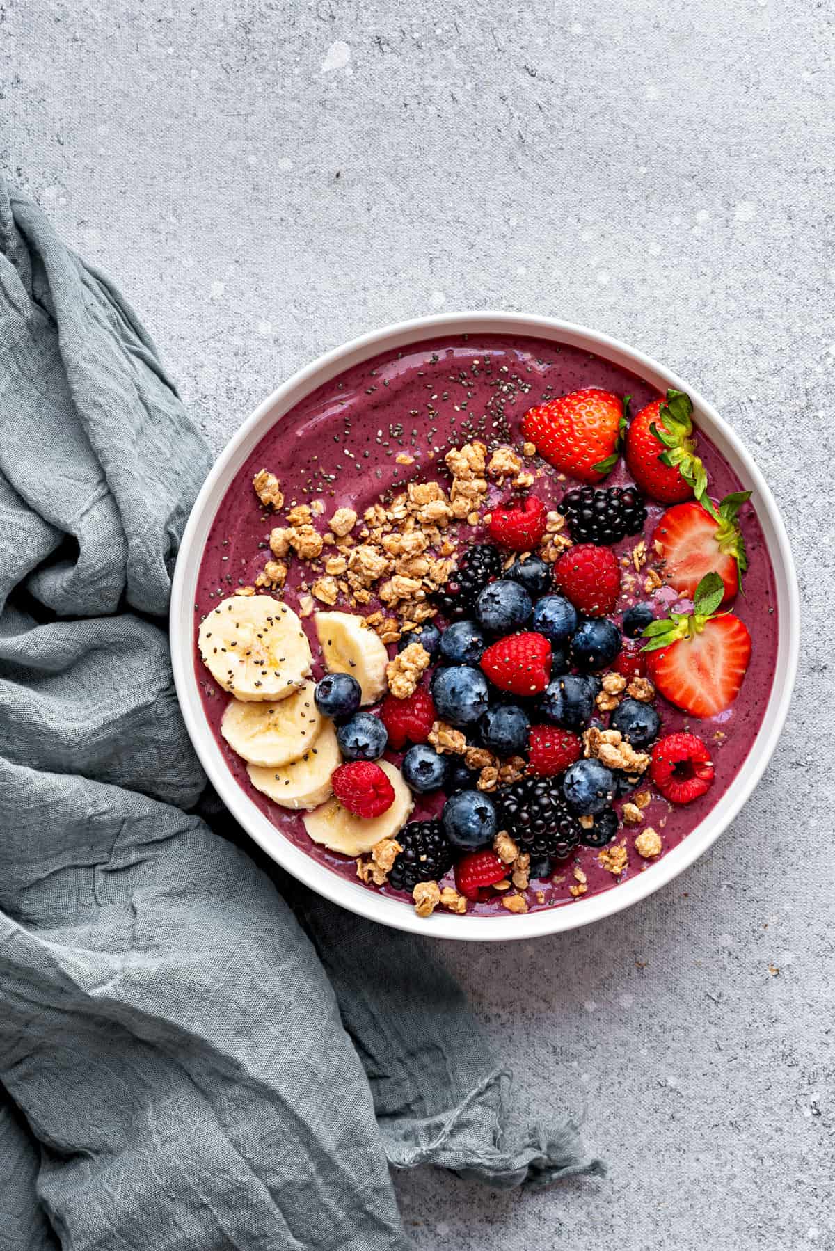 Large white bowl with blended acai berry smoothie, topped with slices of bananas, granola, fresh berries and chia seeds.