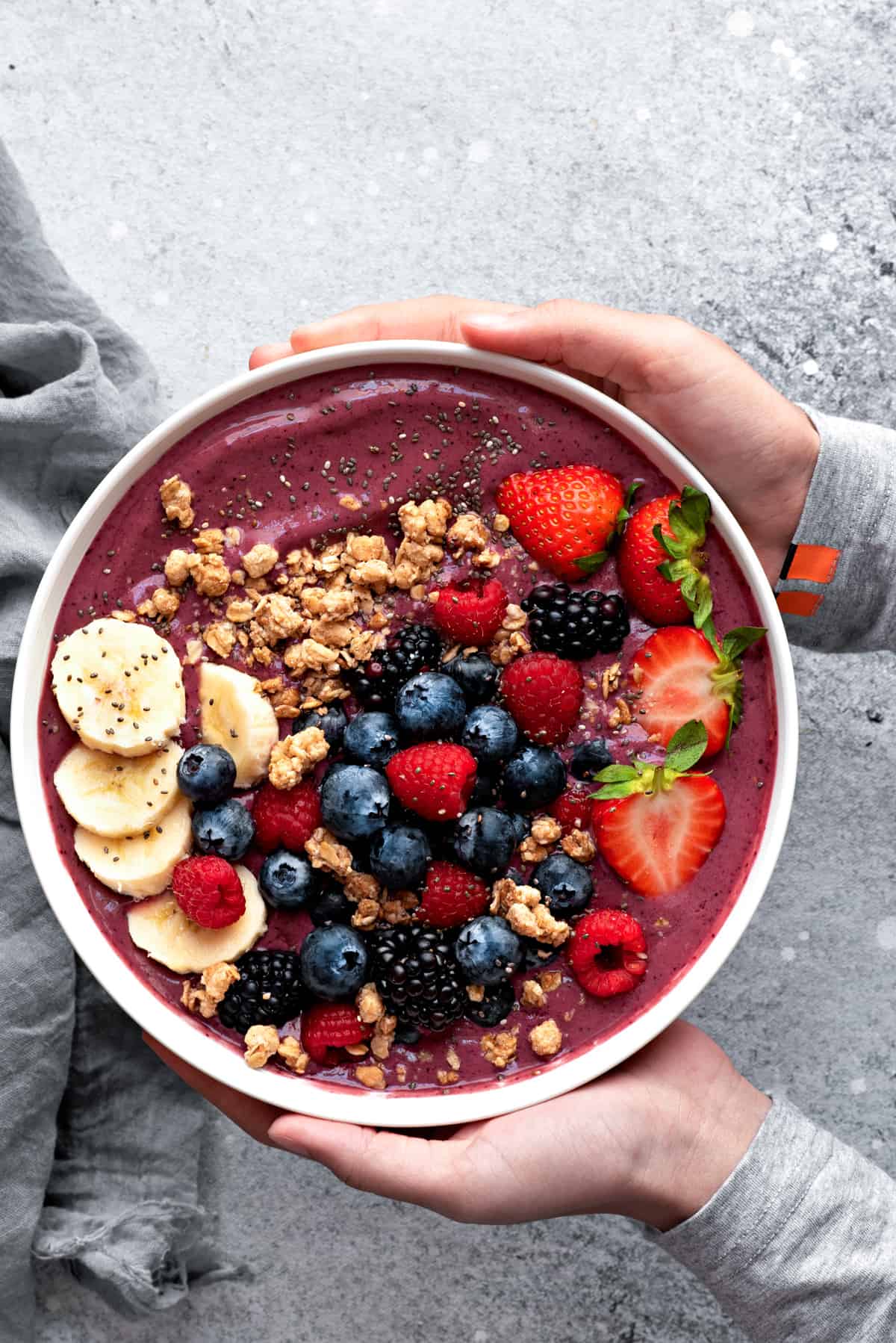 Two hands holding a large white bowl with blended berry Acai smoothie, topped with slices of bananas, granola, fresh blueberries, strawberries, raspberries, and seeds.
