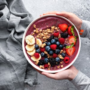 Two hands holding a prepared berry acai bowl, topped with slices of bananas, granola, fresh blueberries, strawberries, raspberries, and chia seeds.