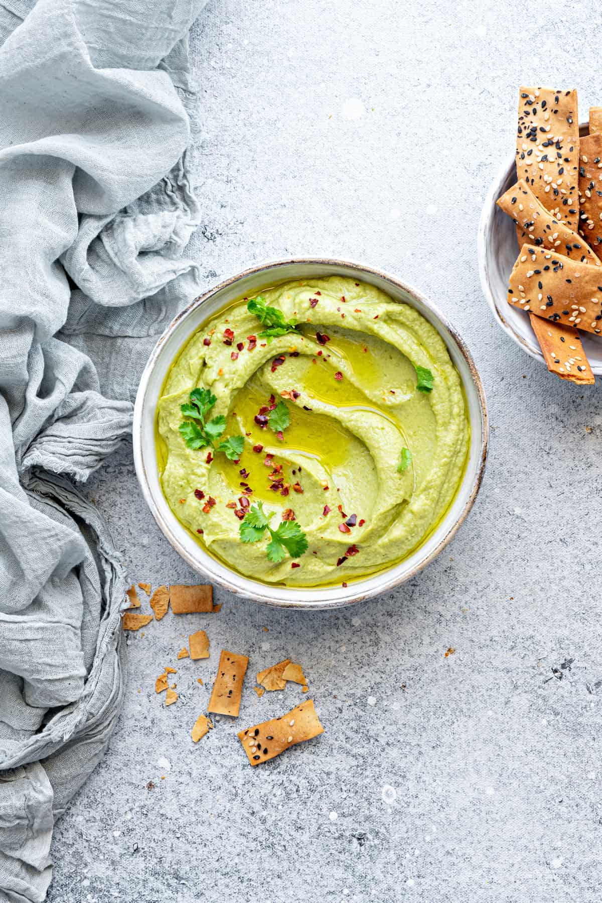 White bowl filled with creamy avocado hummus, topped with fresh cilantro and red pepper flakes. There are pieces of broken cracker scattered to the side.