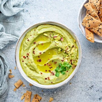 Creamy avocado hummus in white bowl, topped with fresh cilantro, pepper flakes and pistachios.