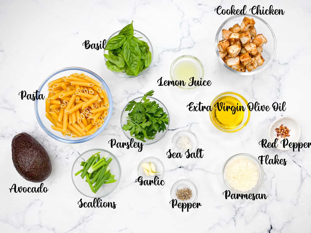 Recipe ingredients each in individual glass bowls: Pasta, fresh basil, fresh parsley, lemon juice, cooked chicken, extra virgin olive oil, red pepper flakes, grated parmesan, salt, pepper, garlic cloves, scallions, and a whole avocado.