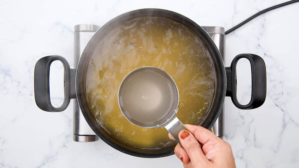 Hand with metal measuring cup, scooping pasta water from the large pot of noodles.