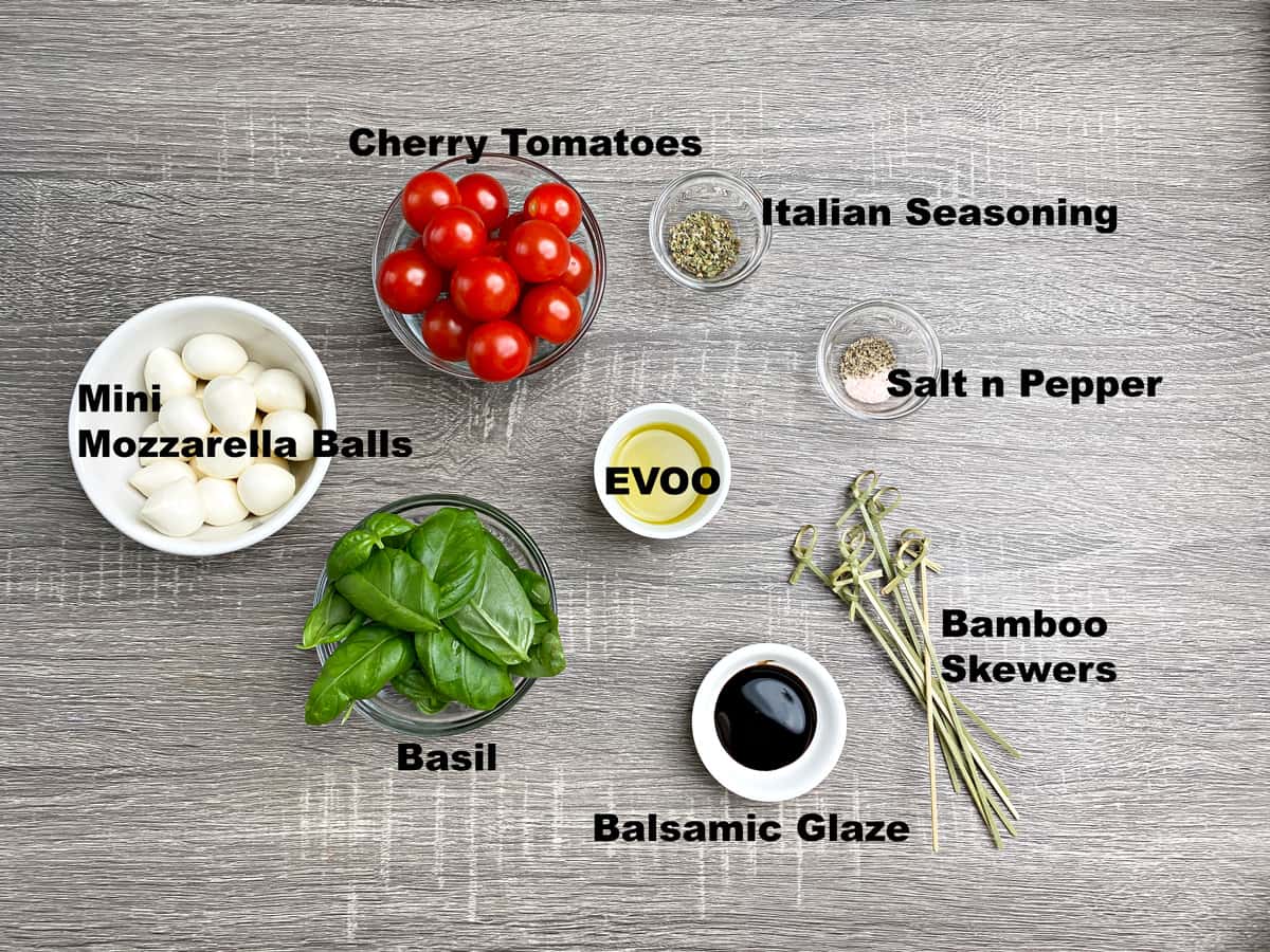 Recipe ingredients, each in a separate dish: Cherry tomatoes, mozzarella cheese balls, basil leaves, evoo, Italian seasoning, salt and pepper, balsamic glaze. Several wood skewers also on the board.