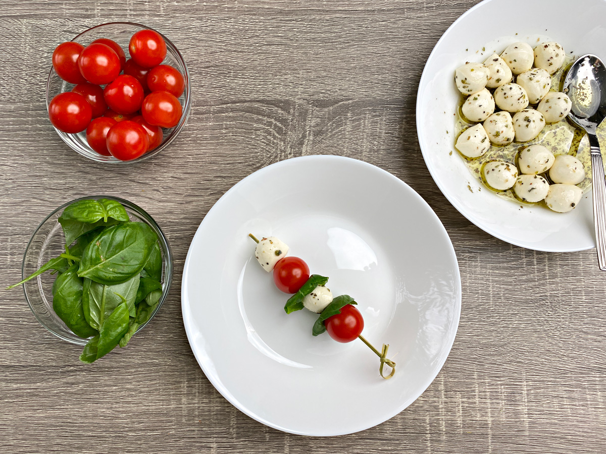 Single Caprese skewer on a white plate: On a skewer is two cherry tomatoes, two mozzarella cheese balls, and two basil leaves.