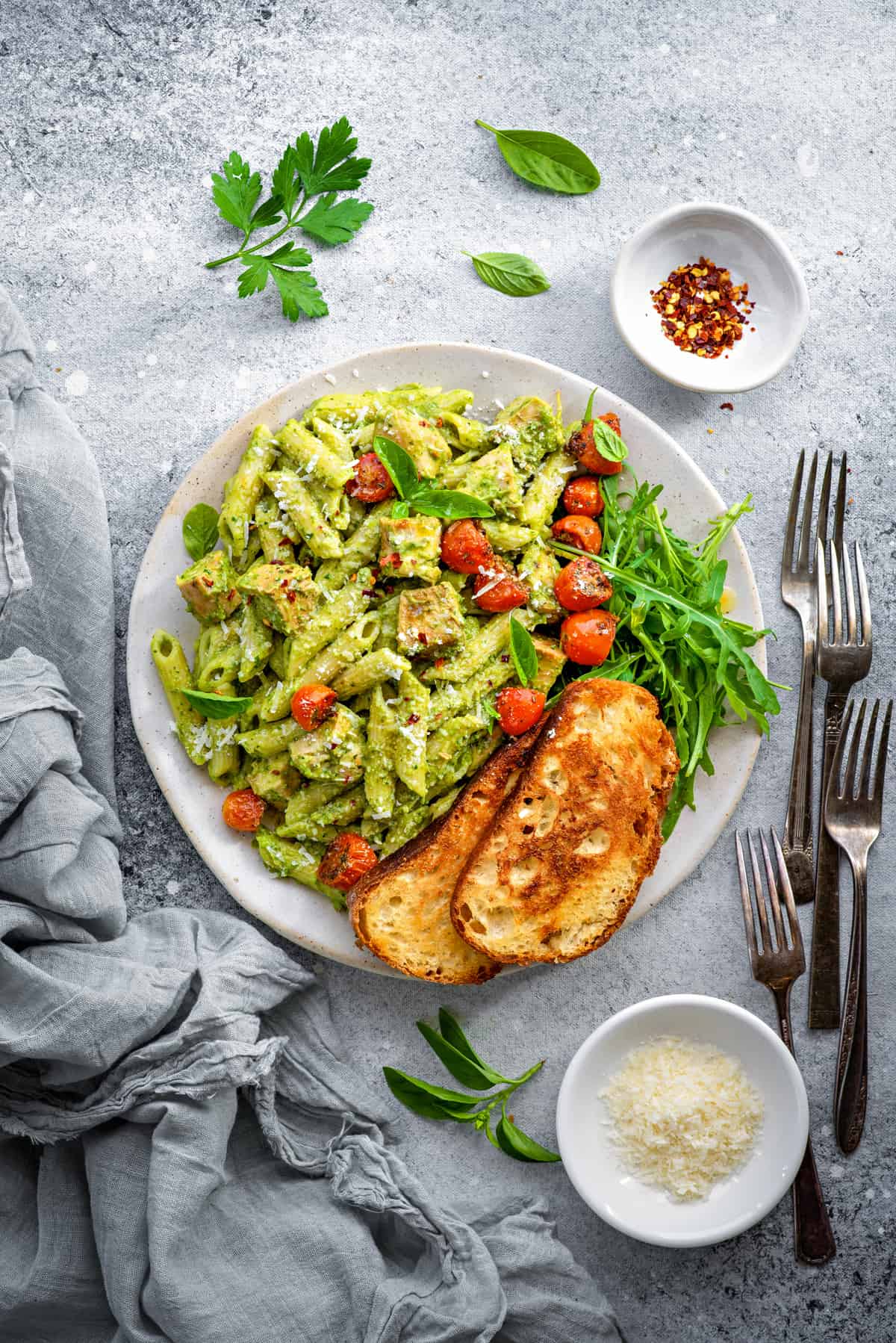 White plate with avocado pasta topped with roasted cherry tomatoes, and two toasted bread slices on the side.