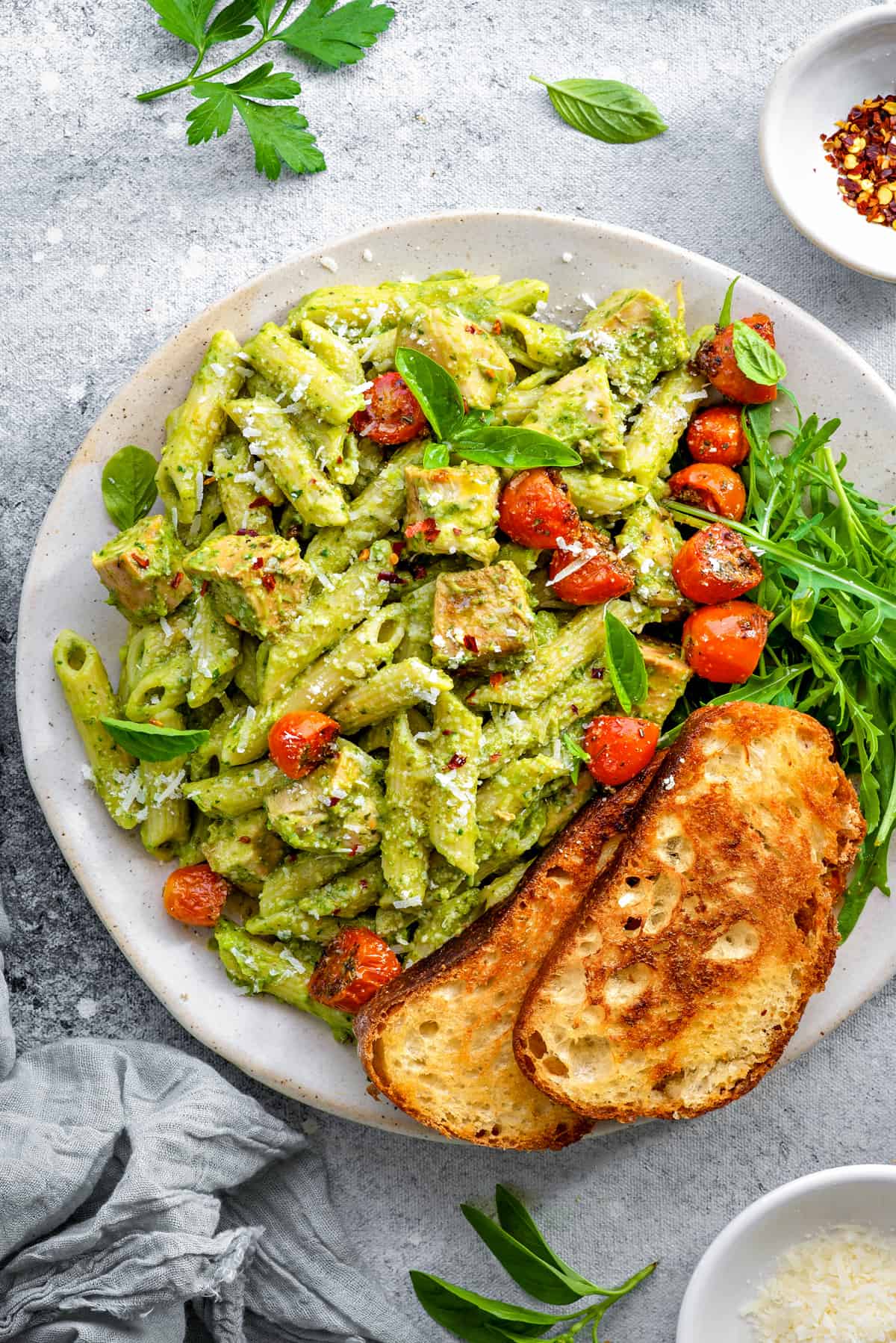 Top down close view white plate with creamy avocado pasta topped with roasted cherry tomatoes, arugula and toasted sourdough.