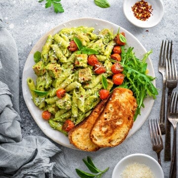 Top down close view white plate with creamy avocado pasta topped with roasted cherry tomatoes, arugula and toasted sourdough.