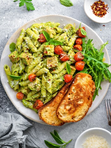 Top down close view white plate with creamy avocado pasta topped with roasted cherry tomatoes, arugula and toasted sourdough.